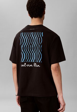 Mees kannab musta t-särki, millel on sinised lainelised jooned ja tekst "calvin klein" seljal, seisab ühevärvilise halli tausta ees.