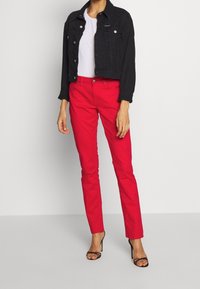 Veste en denim noir avec des boutons, t-shirt blanc et pantalon rouge ajusté. Le modèle porte des escarpins noirs à lanières.