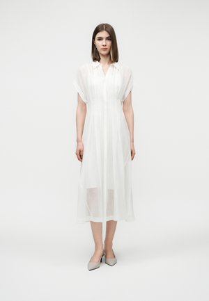 Žena s rovnými hnedými vlasmi má na sebe priehľadné biele midi šaty s krátkym rukávom a sivé lodičky so špicatou špičkou, stojaca pred bielym pozadím.