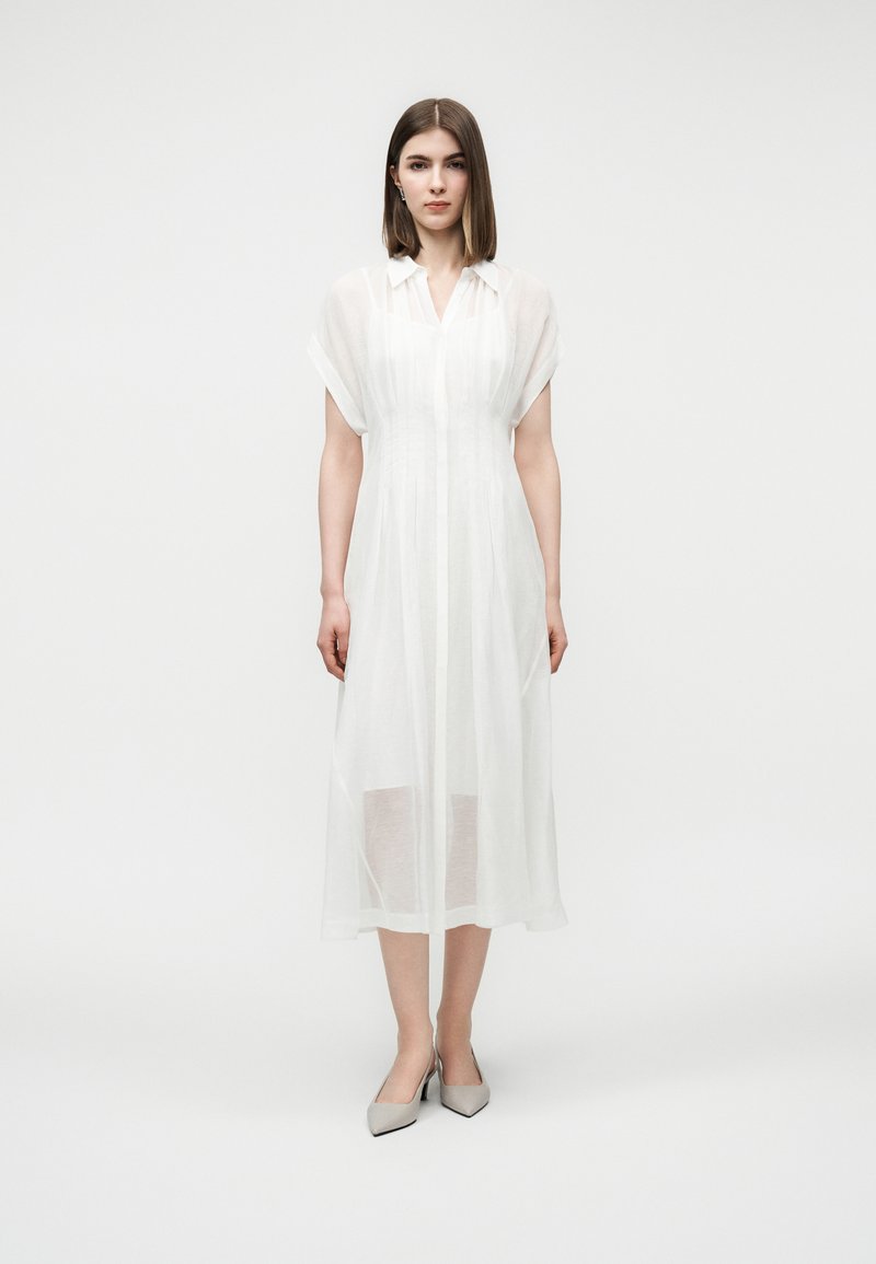 Femme aux cheveux bruns lisses portant une robe midi blanche transparente à manches courtes et des talons gris à bout pointu, debout devant un fond blanc.