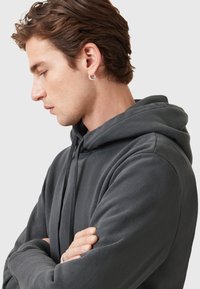 Dunkelgrauer Kapuzenpullover mit weicher Textur, ausgestattet mit einer Fronttasche und einer Kordelzug-Kapuze. Modell mit verschränkten Armen, seitliches Profil sichtbar.