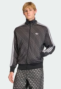 Čierna bundá na zips s vysokým golierom, bielym logom Adidas a tromi bielymi pruhmi pozdĺž rukávov; v kombinácii so zhodnými nohavicami.