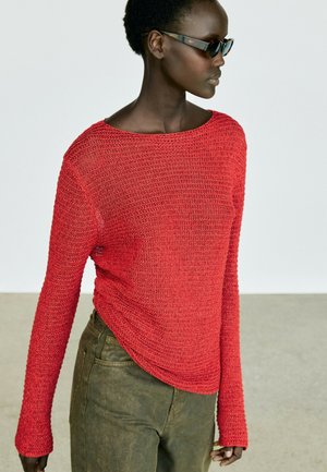 Donna con occhiali da sole scuri, un top rosso lavorato a maglia a maniche lunghe e pantaloni verde oliva, in piedi contro uno sfondo chiaro e uniforme.
