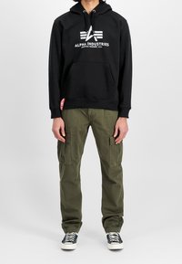 Felpa nera in cotone con logo "ALPHA INDUSTRIES" e tasca a marsupio; abbinata a pantaloni cargo verde oliva, tessuto strutturato e tasche laterali.
