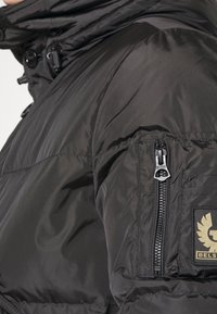 Zwarte gewatteerde jas met een ritszak op de mouw, met een getextureerde afwerking en een logopatch. Het ontwerp heeft een hoge kraag.