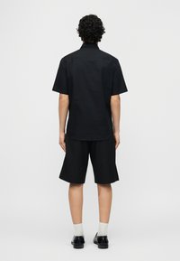 Άνδρας με σγουρά μαλλιά που φοράει μαύρο κοντομάνικο πουκάμισο και μαύρο σορτς μέχρι το γόνατο, στέκεται στραμμένος προς ένα απλό ανοιχτό γκρι φόντο.