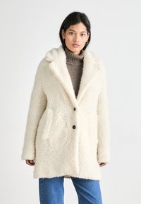 Cream-colored, fluffy coat with a notched collar, black buttons, and two side pockets. Worn over a brown turtleneck and blue jeans.