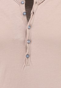 Beige Henley-Shirt mit einer Knopfleiste und fünf blauen Knöpfen, aus texturiertem Stoff und mit einem runden Halsausschnitt. Einfaches, funktionales Design.