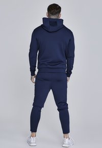 Tuta navy con felpa con cappuccio e pantaloni affusolati. Tessuto morbido con polsini e vita a costine, che evidenzia un design aderente.