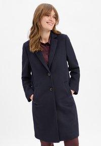Navyblauwe wollen jas met lange mouwen, geknipte kraag, twee voorzakken en twee knopen. Glide textuur en lengte tot halverwege de bovenbenen.