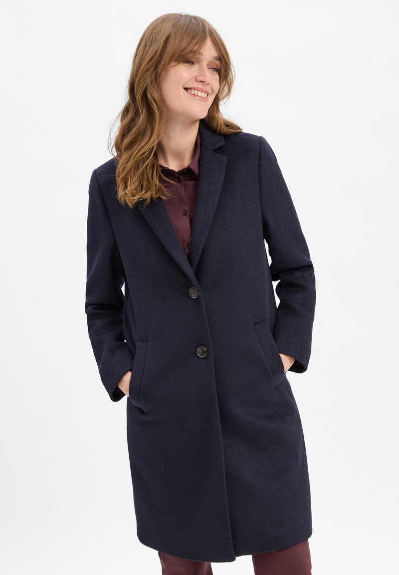 Navyblauwe wollen jas met lange mouwen, geknipte kraag, twee voorzakken en twee knopen. Glide textuur en lengte tot halverwege de bovenbenen.