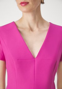 Femme portant une robe rose vif à col en V, manches courtes, et de petites boucles d'oreilles en or, sur un fond clair uni.