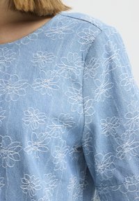 Haut en denim bleu clair avec des motifs floraux blancs brodés sur l'épaule et le haut du dos, porté par une personne aux cheveux blonds courts.