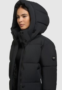 Čierna páperová bunda s veľkou kapucňou, zapínaním na zips a cvočky, vypchávkovaným dizajnom, s logom na ľavom rukáve a hladkou textúrou.