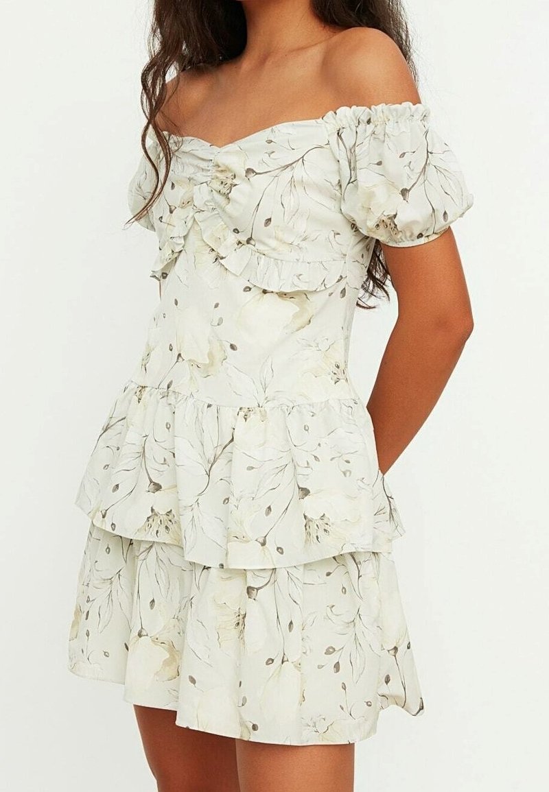 Femme portant une robe blanche épaules dénudées avec manches ballon et motif floral beige subtil, avec un corsage froncé et une jupe à volants.