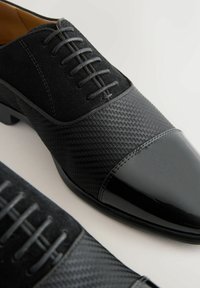 Schwarze Herrenschuhe mit strukturiertem Stoff, Wildlederseiten und glänzenden Lederzehen, geschnürt mit schwarzen Schnürsenkeln vor einem weißen Hintergrund.