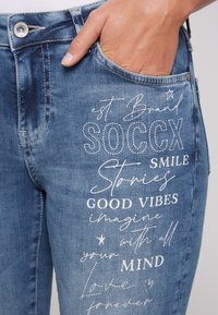 Vaqueros de denim en un lavado azul claro con costuras visibles. Presentan texto impreso en blanco, incluyendo "SOCCX" y varias frases, en la pierna izquierda.
