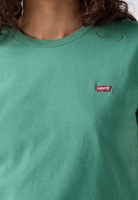 Gros plan d'une personne portant un t-shirt vert à col rond avec un petit écusson rouge du logo Levi's sur la poitrine gauche.