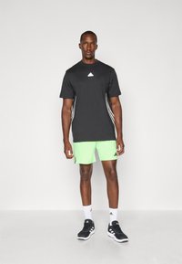 T-shirt de sport noire avec logo blanc, associé à un short vert vif. La tenue présente des rayures blanches sur les côtés et des chaussures de sport noires.