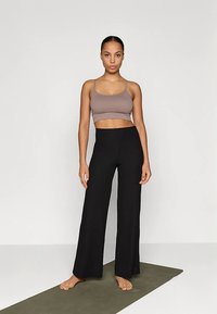 Brown cropped tank top and black wide-leg ribbed pants. Model stands on a green mat, barefoot, against a plain white background.
