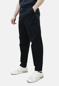 Pantaloni neri realizzati in tessuto leggero, con vestibilità slim, tasche laterali e chiusura con bottone, abbinati a scarpe sportive bianche.