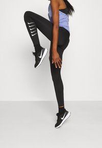 Czarne legginsy sportowe z białym logo Nike i paskami, połączone z fioletową bluzką bez rękawów oraz czarnymi butami sportowymi z białymi akcentami.