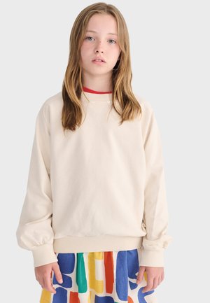 Jeune personne aux cheveux longs portant un sweat-shirt crème et une jupe à motifs colorés, debout devant un fond uni.