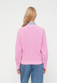 Sweater rosa em malha com um design texturizado, punhos e bainha canelados. Apresenta um detalhe de colarinho azul em camadas. Usado com jeans de denim largos.