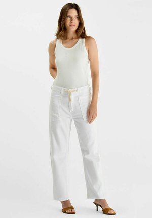 Vrouw staand, gekleed in een wit mouwloos topje, witte broek met trekkoord en zakken aan de voorkant, en bruine open-toe sandalen met hak.