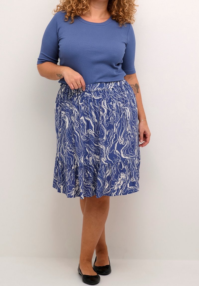Femme portant un haut bleu à manches courtes et une jupe bleu et blanche à motifs longueur genou avec des ballerines noires, debout devant un fond uni.