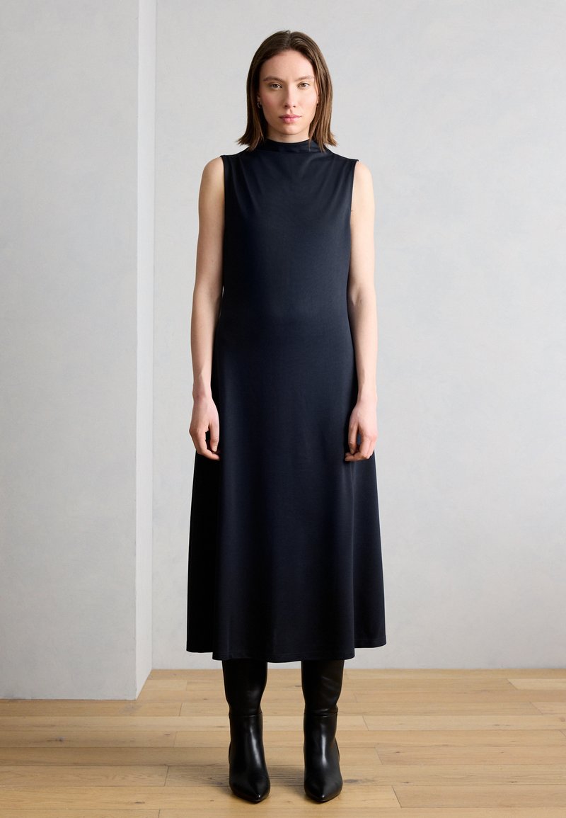 Femme debout à l'intérieur portant une robe midi noire sans manches et des bottes noires montantes jusqu'aux genoux, devant un mur gris clair uni.