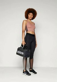 Naine roosas lõhikutopis ja mustades säärises, millel on läbipaistvad paneelid, hoiab musta Adidas'i kottu, seistes ühevärvilise tausta ees.