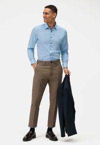 Chemise bleu clair à boutons noirs, associée à un pantalon tailleur marron. Un homme tient un blazer bleu foncé avec une texture lisse.