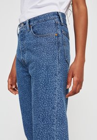Blå denimjeans med mönster av leopardtryck, hög midja, rak bendesign och diskreta sömadetaljer, visade från sidan med handen vilande.