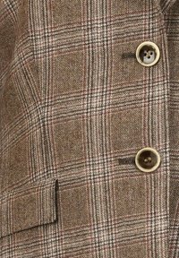 Hnědý kostkovaný blazer s texturovanou látkou. Má zlaté knoflíky a klopovou kapsu na levé straně, s pevně zakončeným střihem.