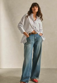 Chemise blanche surdimensionnée à boutons, jeans large bleu clair, ceinture en imprimé léopard, ballerines rouges. Tissu doux, coupe décontractée et design casual.