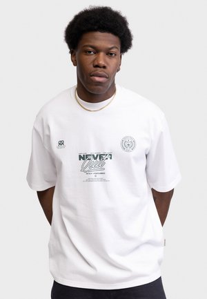 Uomo con afro corto che indossa una t-shirt bianca con grafica e una catena d'oro al collo, in piedi davanti a uno sfondo chiaro e semplice.