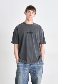 Camiseta gris de gran tamaño, hecha de una tela suave, con un escote redondo y un estampado de logo negro en el frente. Combinada con jeans azul desgastados.