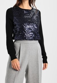 Kobieta w czarnej bluzce z długim rękawem pokrytej błyszczącymi geometrycznymi cekinami oraz w szarych, szerokich spodniach z wysokim stanem, stoi z rękami w luźnej pozycji.