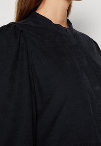 Femme portant une chemise en velours côtelé noire avec un col montant et une fermeture à boutons, montrant les détails des épaules et du haut de la poitrine.