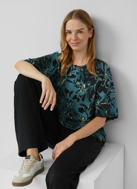 Blouse florale teal avec des accents noirs, manches courtes et col rond ; associée à un pantalon noir et des baskets de couleur claire.