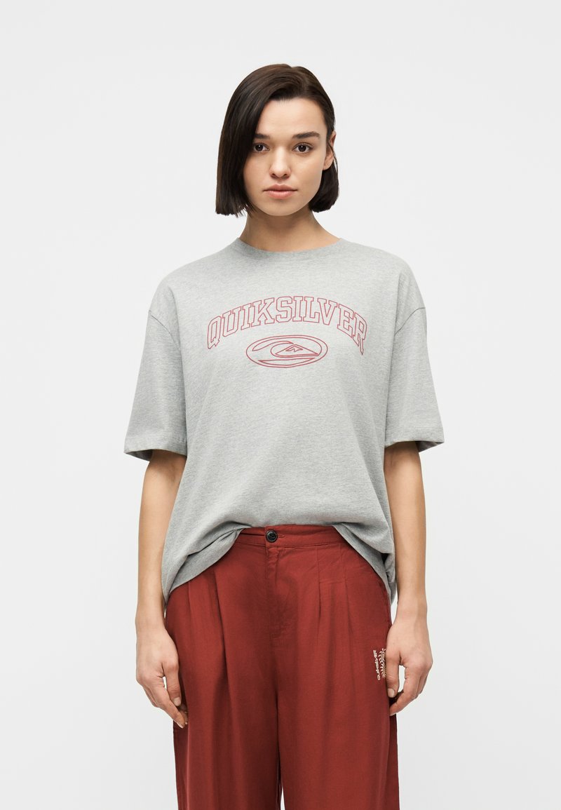 Γυναίκα με κοντά σκούρα μαλλιά, φορώντας μια φαρδιά γκρι μπλούζα Quiksilver που έχει μπει μέσα σε ψηλόμεσα παντελόνια σε σκουροκόκκινο χρώμα, στέκεται μπροστά σε ένα απλό φόντο.