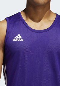 Gros plan d'une personne portant un maillot de sport sans manches Adidas violet avec logo blanc et bandes latérales.