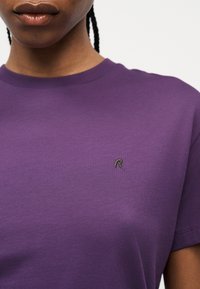 Gros plan d'une personne portant un t-shirt violet à col rond avec un petit logo "R" brodé sur le côté gauche de la poitrine.