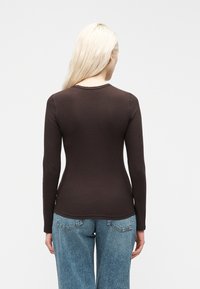 Γυναίκα με μακριά ξανθά μαλλιά που φοράει εφαρμοστή σκούρα καφέ μπλούζα με μακριά μανίκια και μπλε τζιν, φαίνεται από πίσω με φόντο απλό.