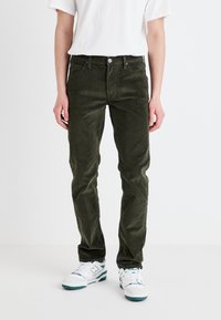 Tumši zaļi korduroja bikses ar taisnu piegriezumu, ar pogu un rāvējslēdzēju, piecas kabatas, apvienotas ar baltām sporta apaviem.