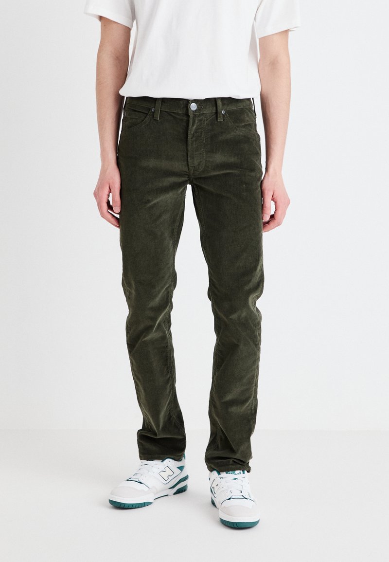 Tumši zaļi korduroja bikses ar taisnu piegriezumu, ar pogu un rāvējslēdzēju, piecas kabatas, apvienotas ar baltām sporta apaviem.