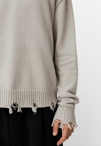 Heller grauer Strickpullover mit geripptem Saum und Bündchen; verfügt über ausfransende Kanten und strukturierten Stoff. Wird über schwarzen Hosen getragen.