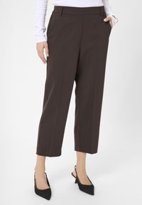 Braune maßgeschneiderte Hose mit seitlichen Taschen, verkürzter Länge, Falten auf der Vorderseite, glatte Textur, kombiniert mit schwarzen Slingback-Schuhen mit spitzem Zehenbereich.