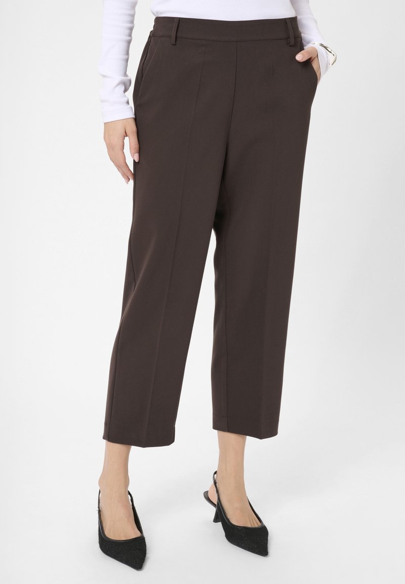 Braune maßgeschneiderte Hose mit seitlichen Taschen, verkürzter Länge, Falten auf der Vorderseite, glatte Textur, kombiniert mit schwarzen Slingback-Schuhen mit spitzem Zehenbereich.